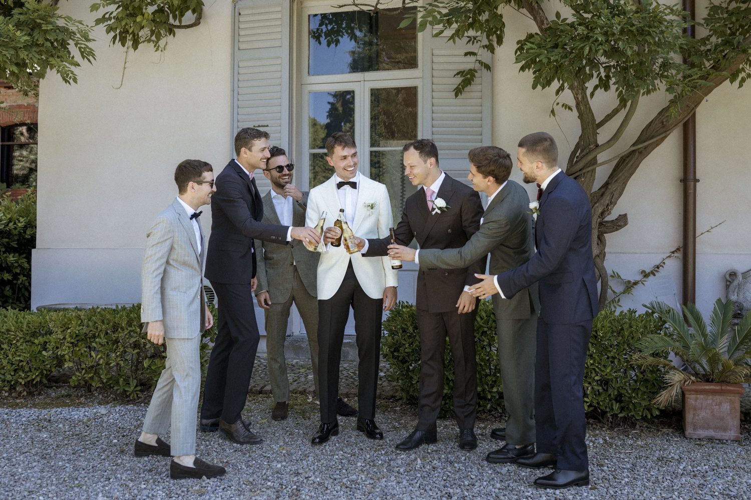 The groom in a white dinner jacket toasting with beers with his groomsmen on the gravel in front of the villa