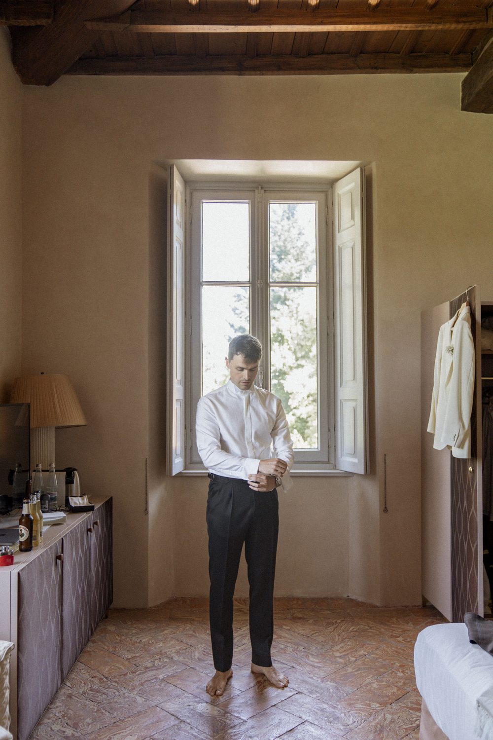 The groom fastening his cuff by the shuttered window of a suite, his white dinner jacket hanging ready on the wardrobe under the beamed ceiling