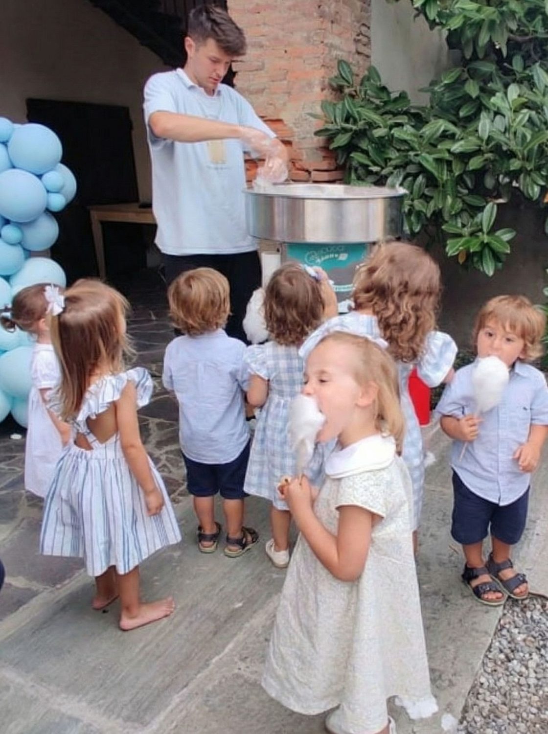 Children in pale blue eating candy floss around the machine in the courtyard, next to a balloon arch