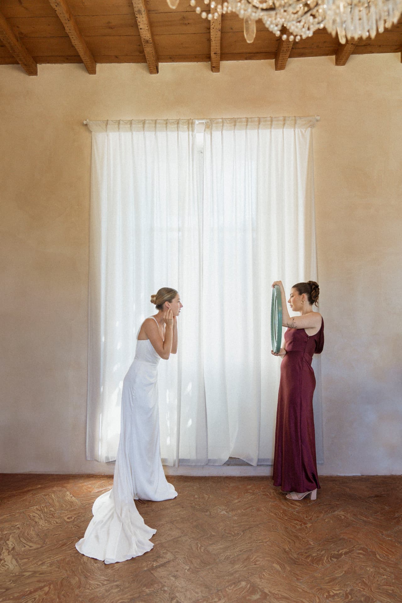 The bride fastening an earring in front of a tall curtained window, her train trailing on the terracotta floor, as a bridesmaid in a burgundy dress holds up a small green mirror under the beamed ceiling