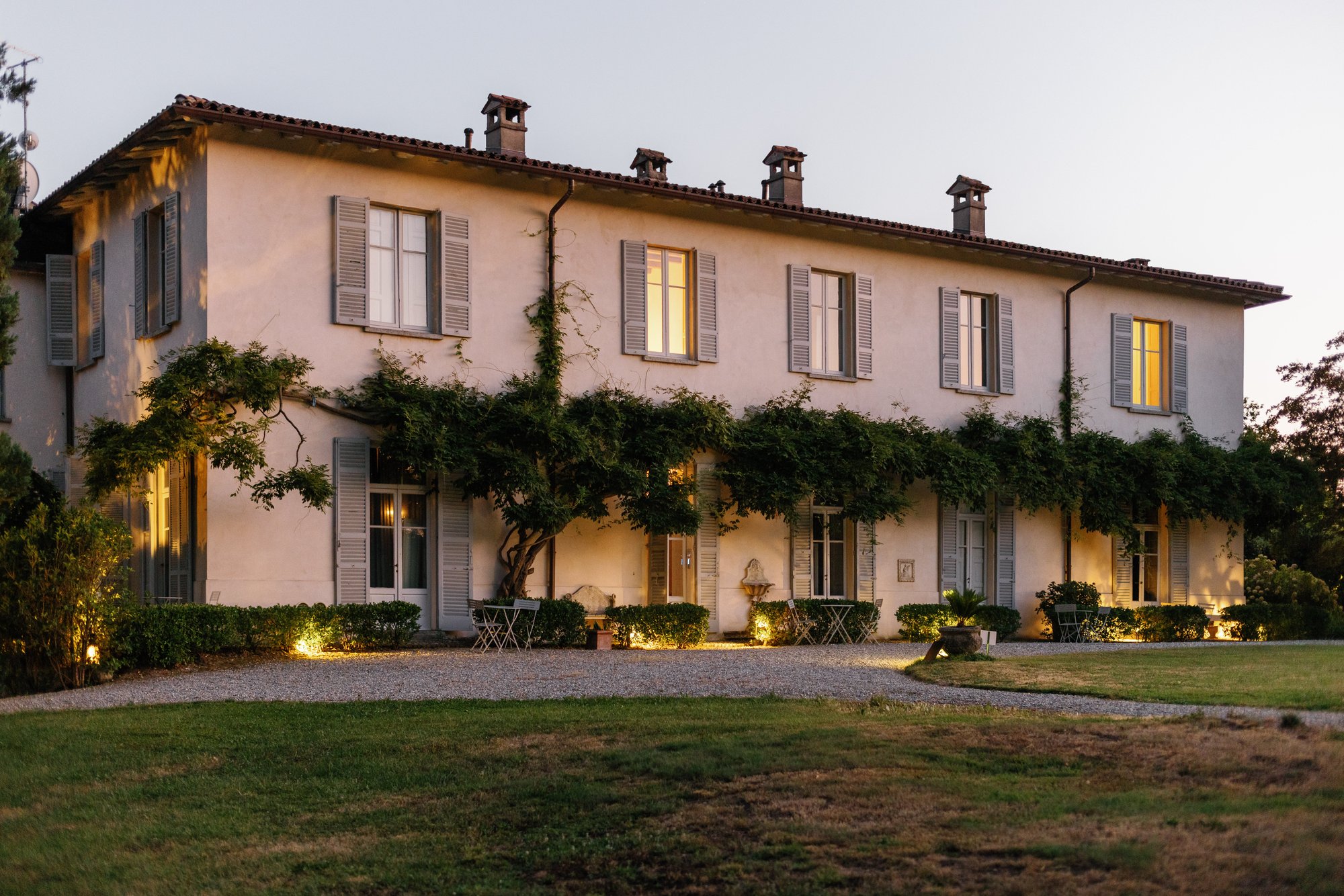 The long villa facade at sunset, with grey shutters, a wisteria trained along the wall and lit windows above the lawn