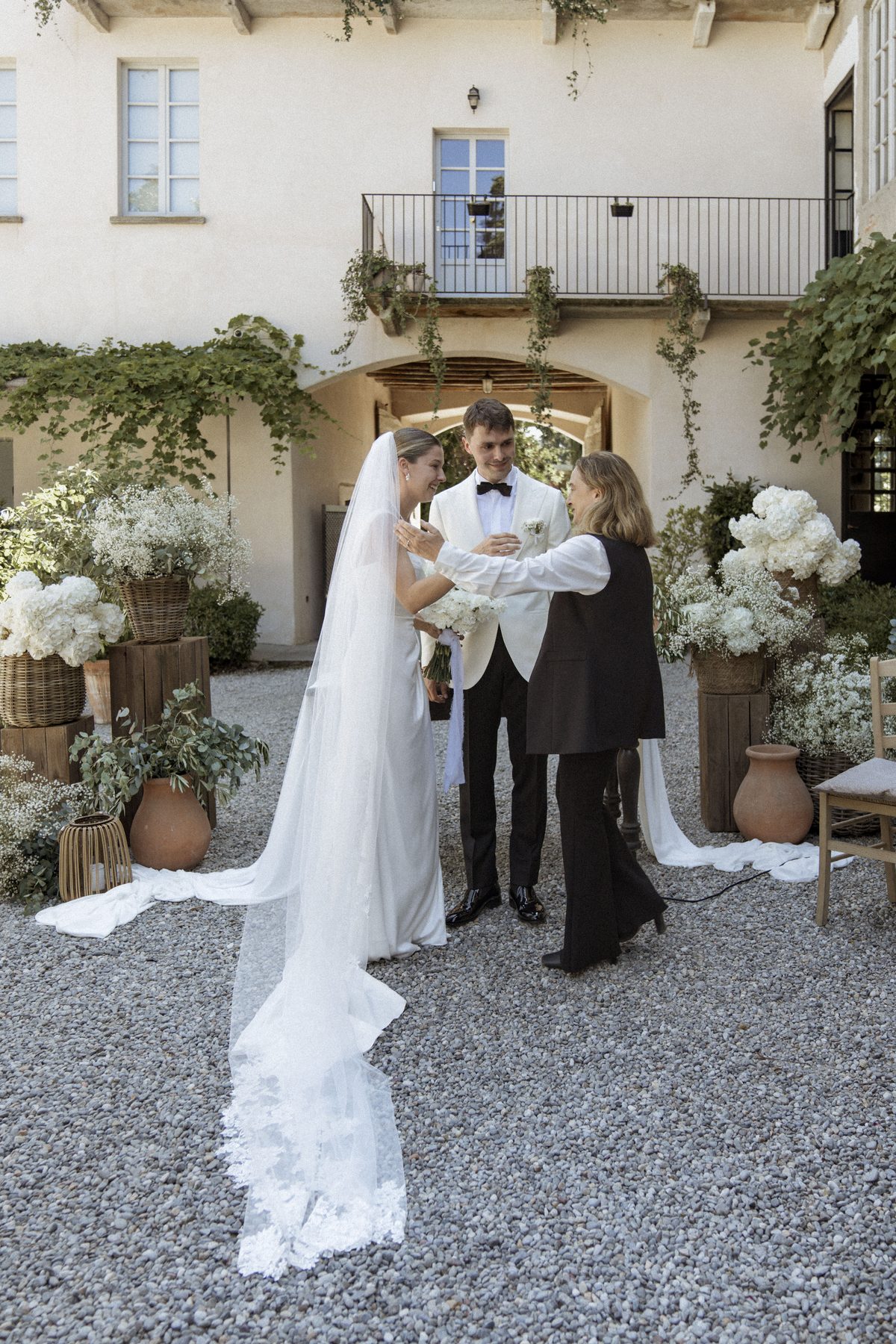 The bride in a long veil and the groom in a white dinner jacket facing the celebrant on the gravel, between baskets of white hydrangeas and gypsophila