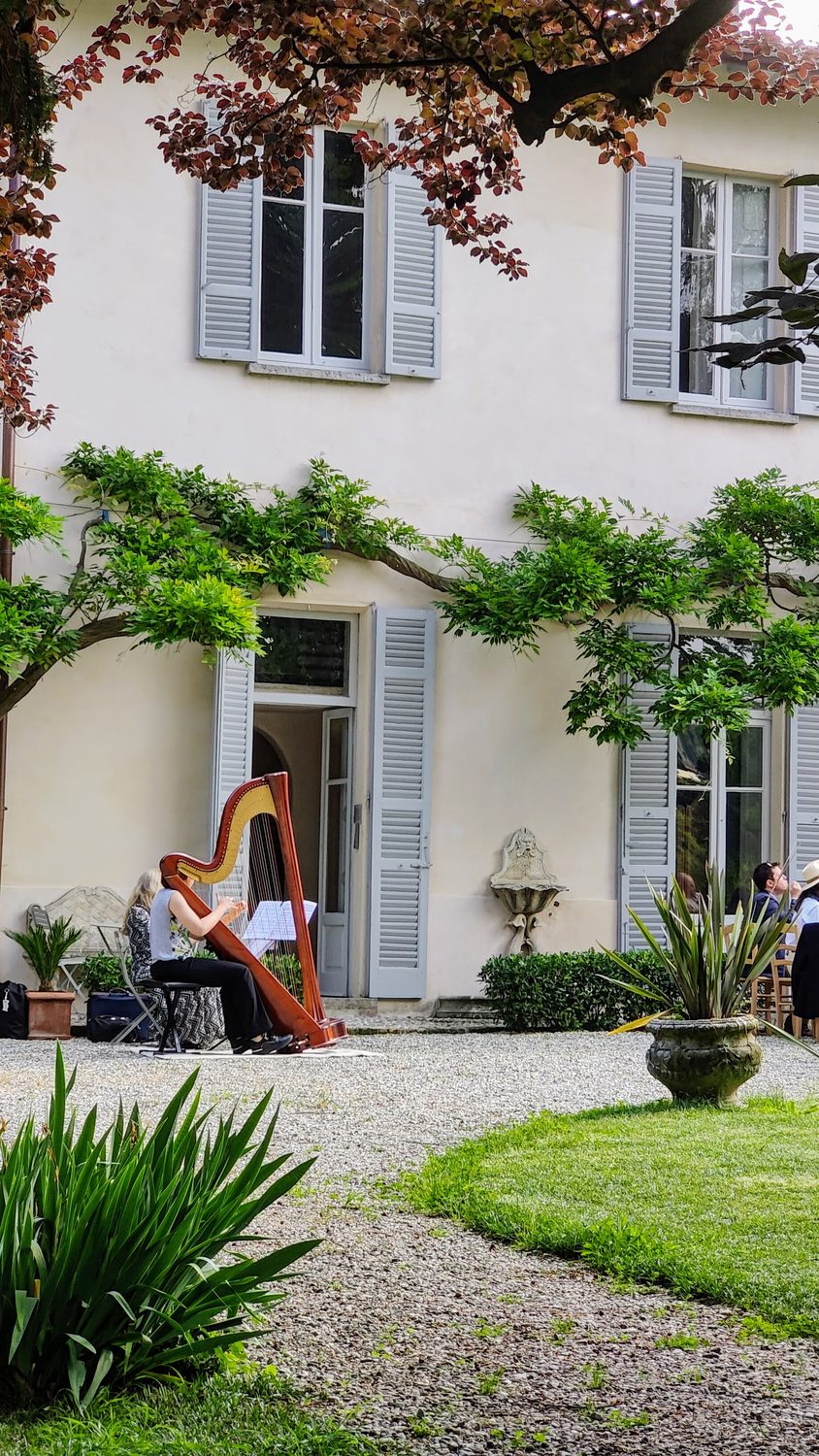 A harpist playing in front of the villa, under the wisteria trained along the facade, with guests seated in the shade on the gravel
