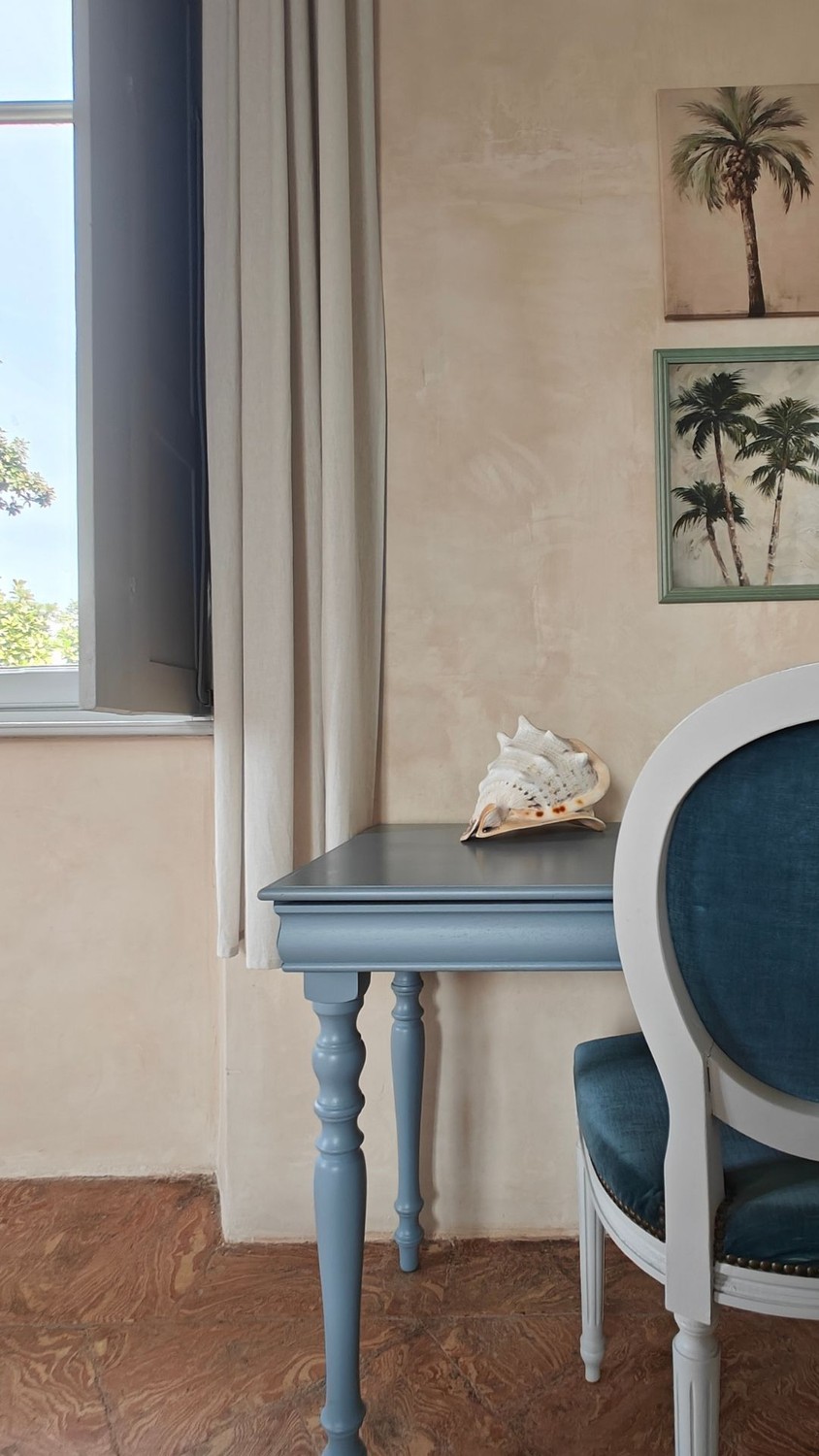 A corner of a suite: a pale blue writing table by the shuttered window, a blue velvet chair and framed palm prints on the plaster wall