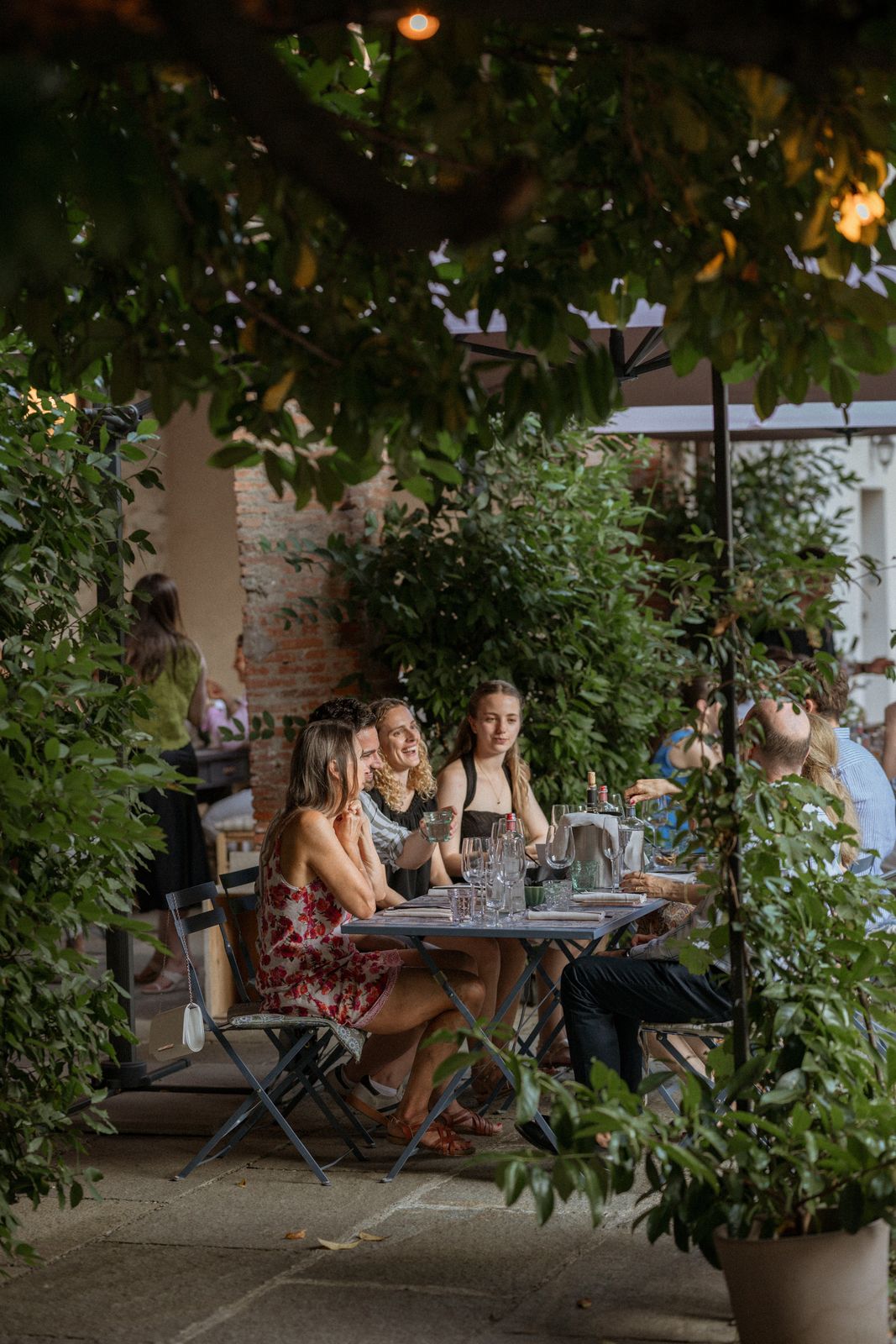 Guests laughing and talking around a small table in the greenery of the courtyard garden, wine glasses and carafes on the tablecloth