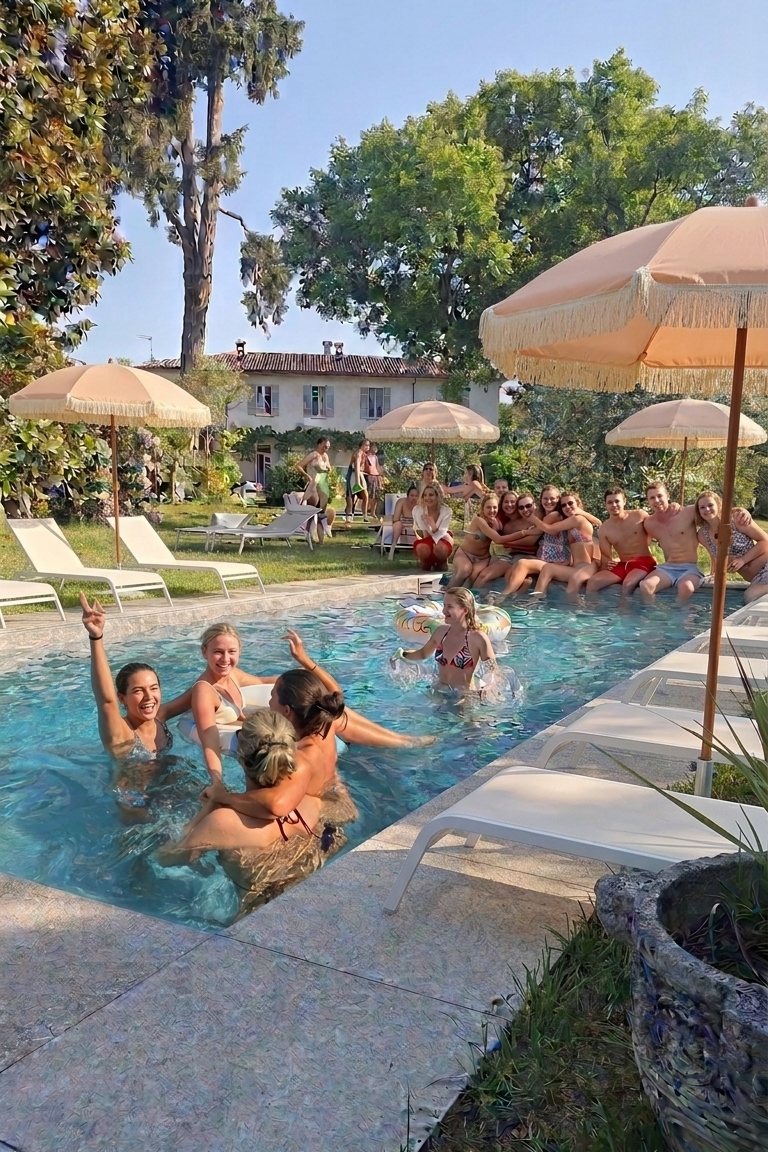 Wedding guests in the swimming pool in the park, laughing in the water and sitting along the edge, with peach fringed parasols and sun loungers on the lawn and the villa behind
