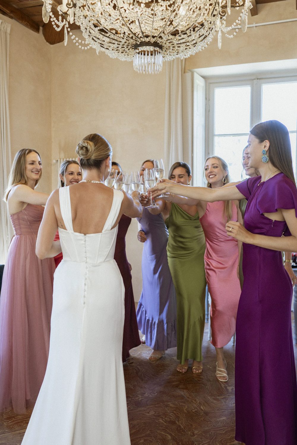 The bride in her white dress raising a glass with her bridesmaids in coloured silk dresses under the crystal chandelier of the villa