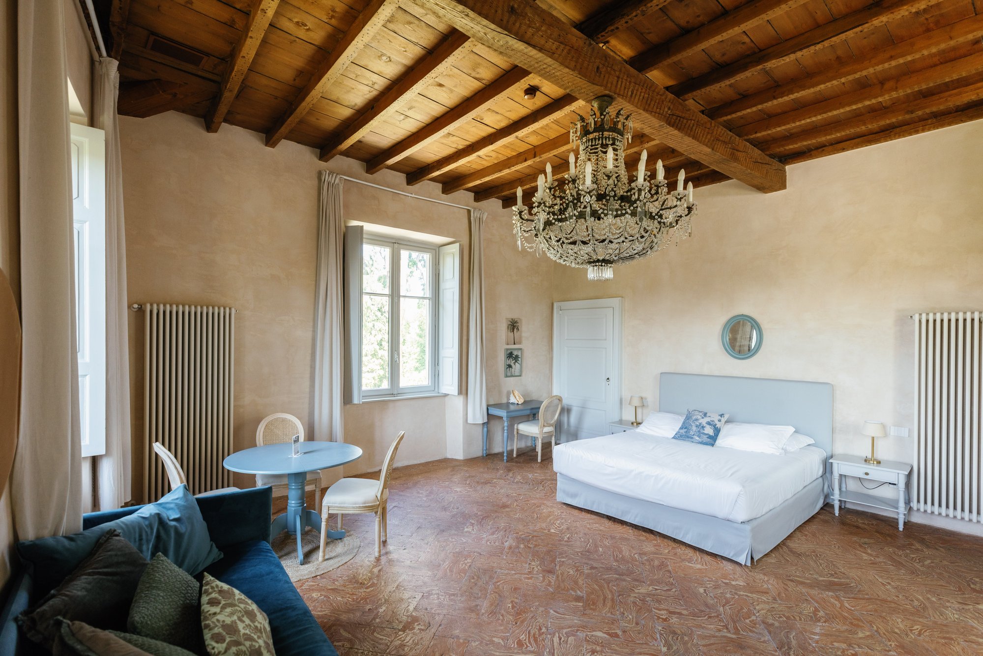 A suite with an exposed wooden beam ceiling, crystal chandelier, terracotta floor, white-linen bed and a blue writing table by the window