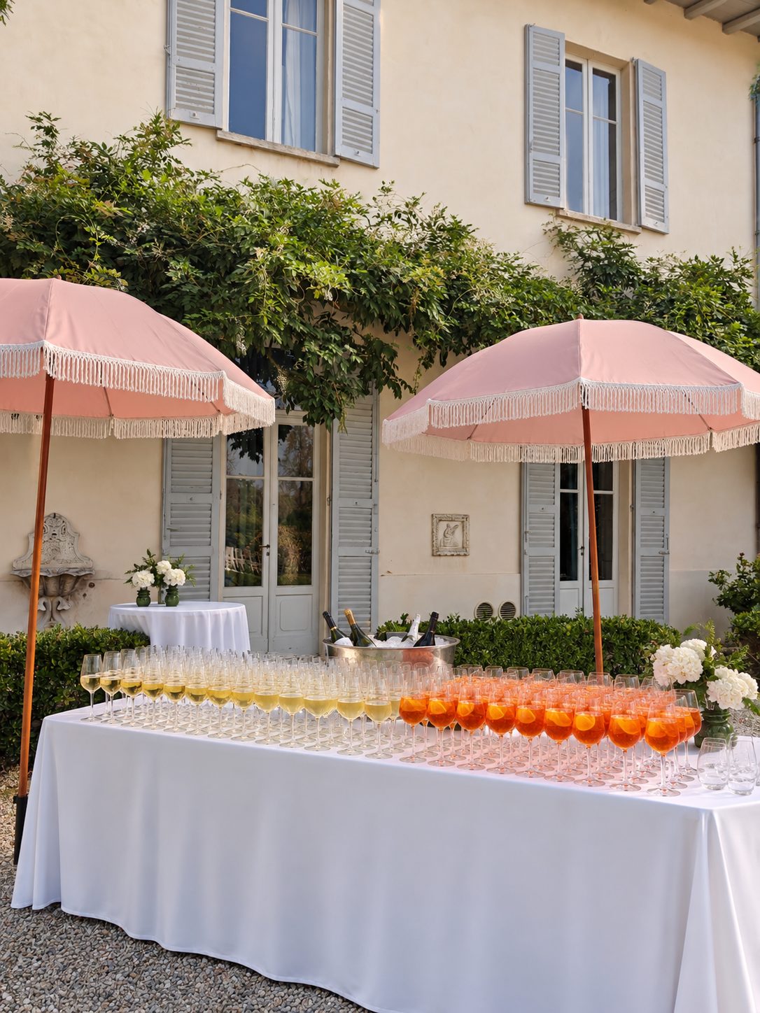 The aperitivo bar in front of the villa: a long white table of prosecco and spritz glasses between two pink fringed parasols, with the shuttered facade and climbing wisteria behind