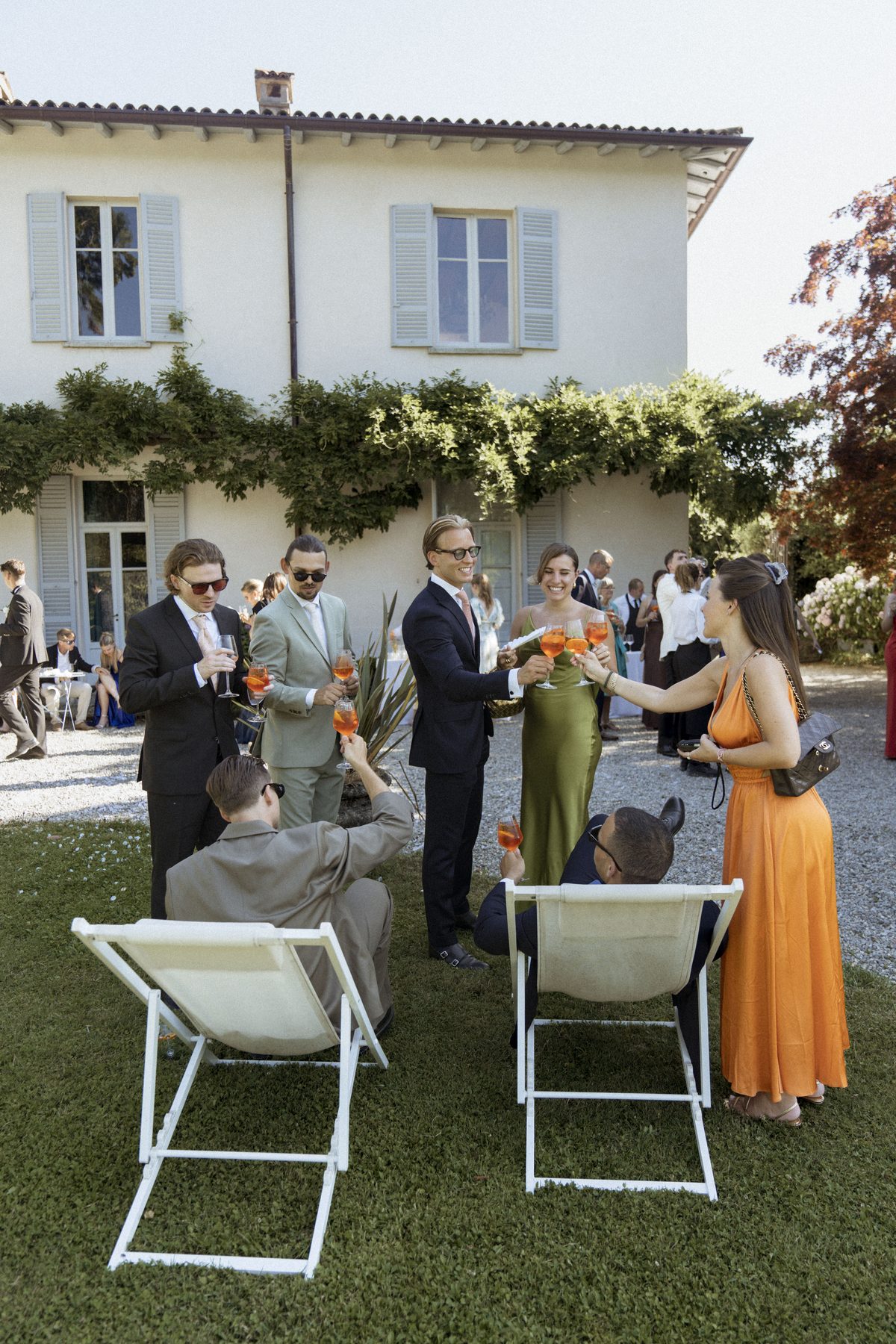 Wedding guests toasting with Aperol spritz on the lawn in front of the villa, between white deckchairs, in green and orange silk dresses and summer suits