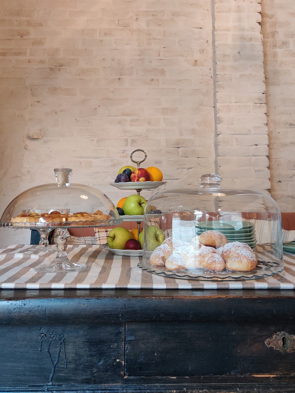 The breakfast table against a whitewashed brick wall: croissants dusted with sugar under a glass dome, a fruit tart on a glass stand and a tiered stand of apples, plums and oranges on a striped linen runner over an old painted chest