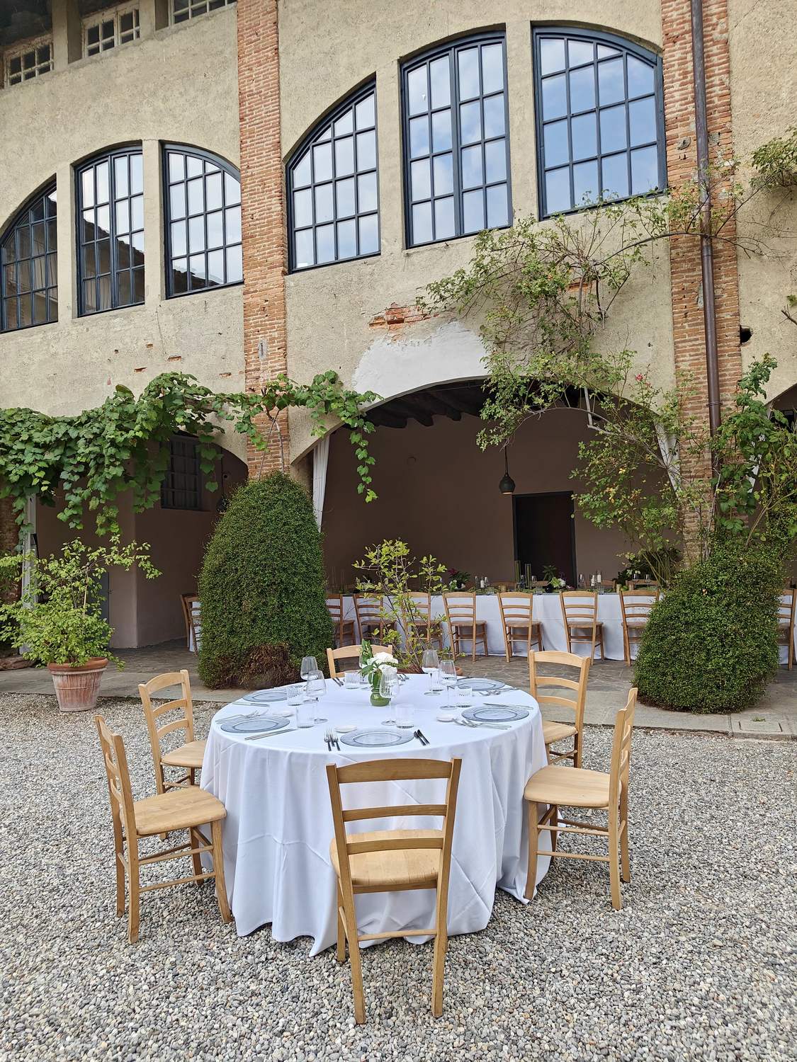 A round table laid with white linen and wooden chairs on the gravel of the courtyard, with the long tables under the portico and the climbing vines behind