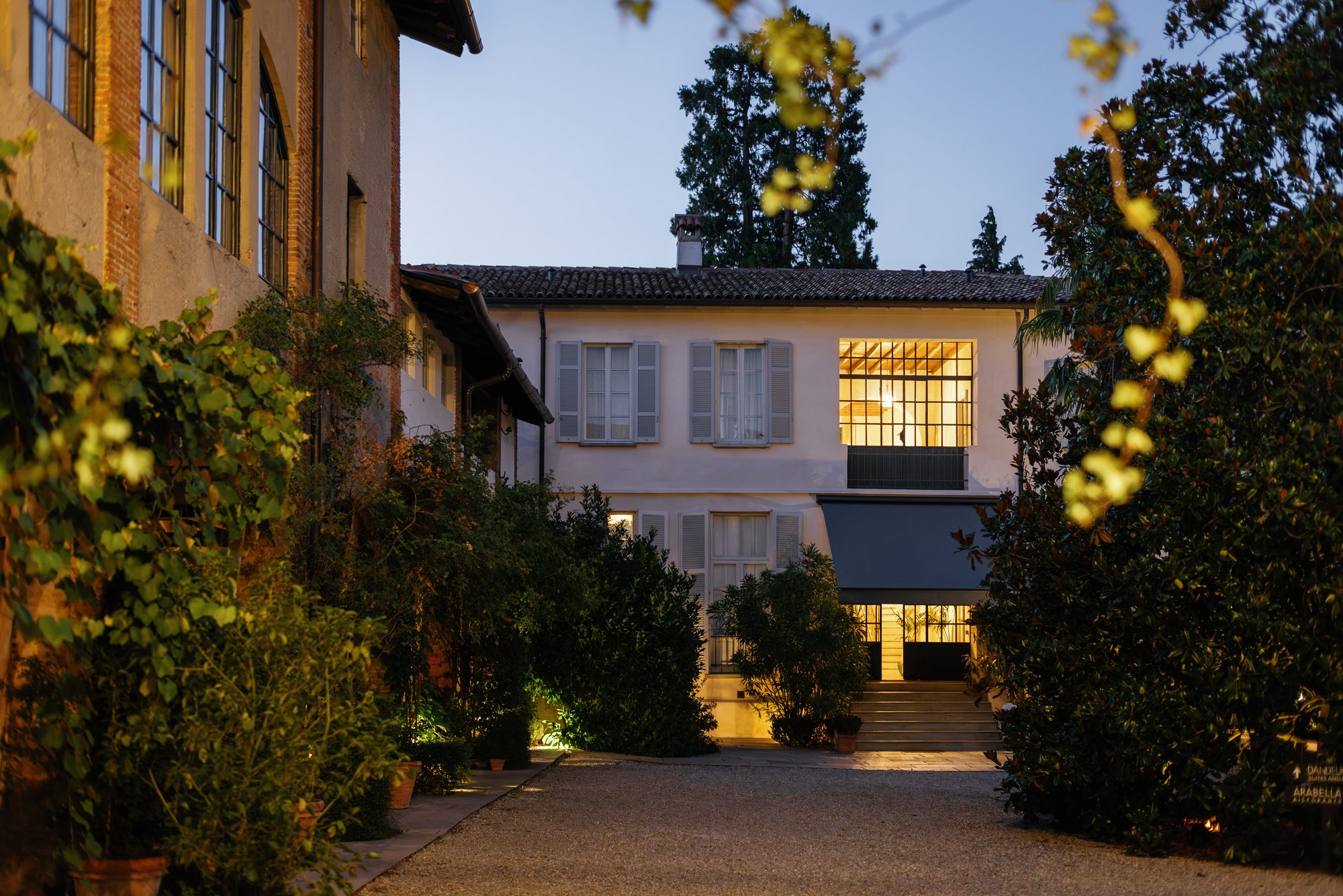 The lit entrance of the estate at dusk, seen from the gravel drive between old brick walls and greenery