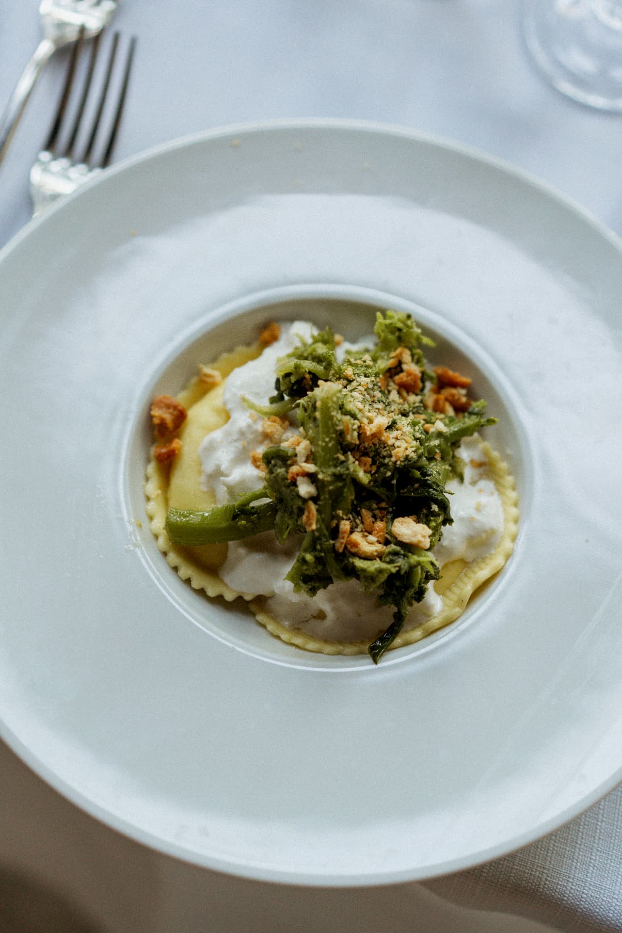 A plate of ravioli with stracciatella, broccoli rabe and toasted crumbs on a white tablecloth beside a silver fork