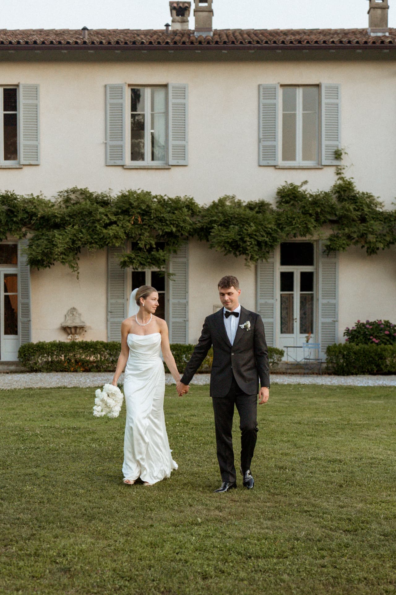 The bride and groom walking hand in hand across the lawn in front of the villa facade at dusk, her bouquet of white flowers in hand