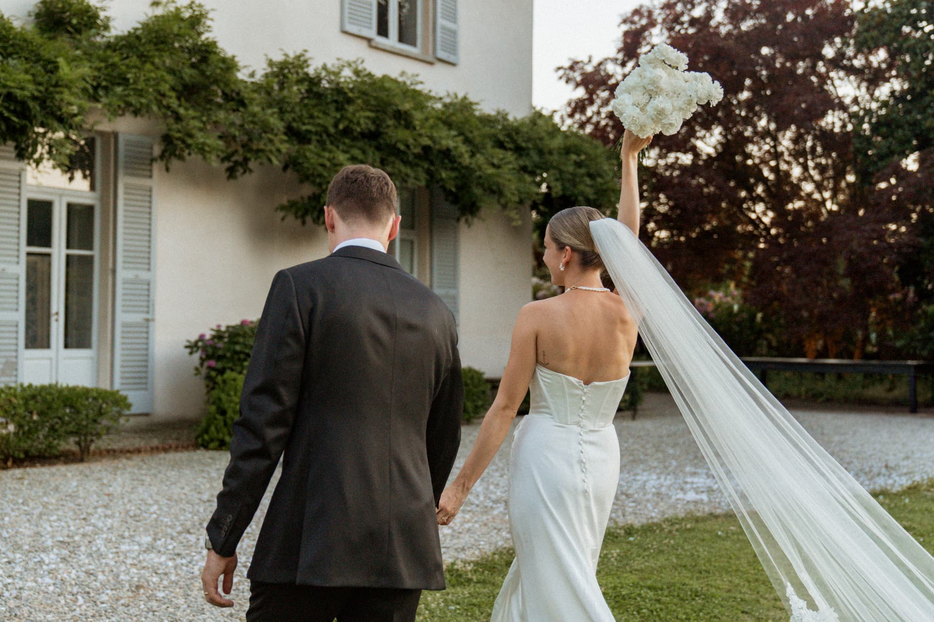 The bride and groom walking hand in hand across the gravel in front of the villa, her long veil trailing and her bouquet of white flowers raised