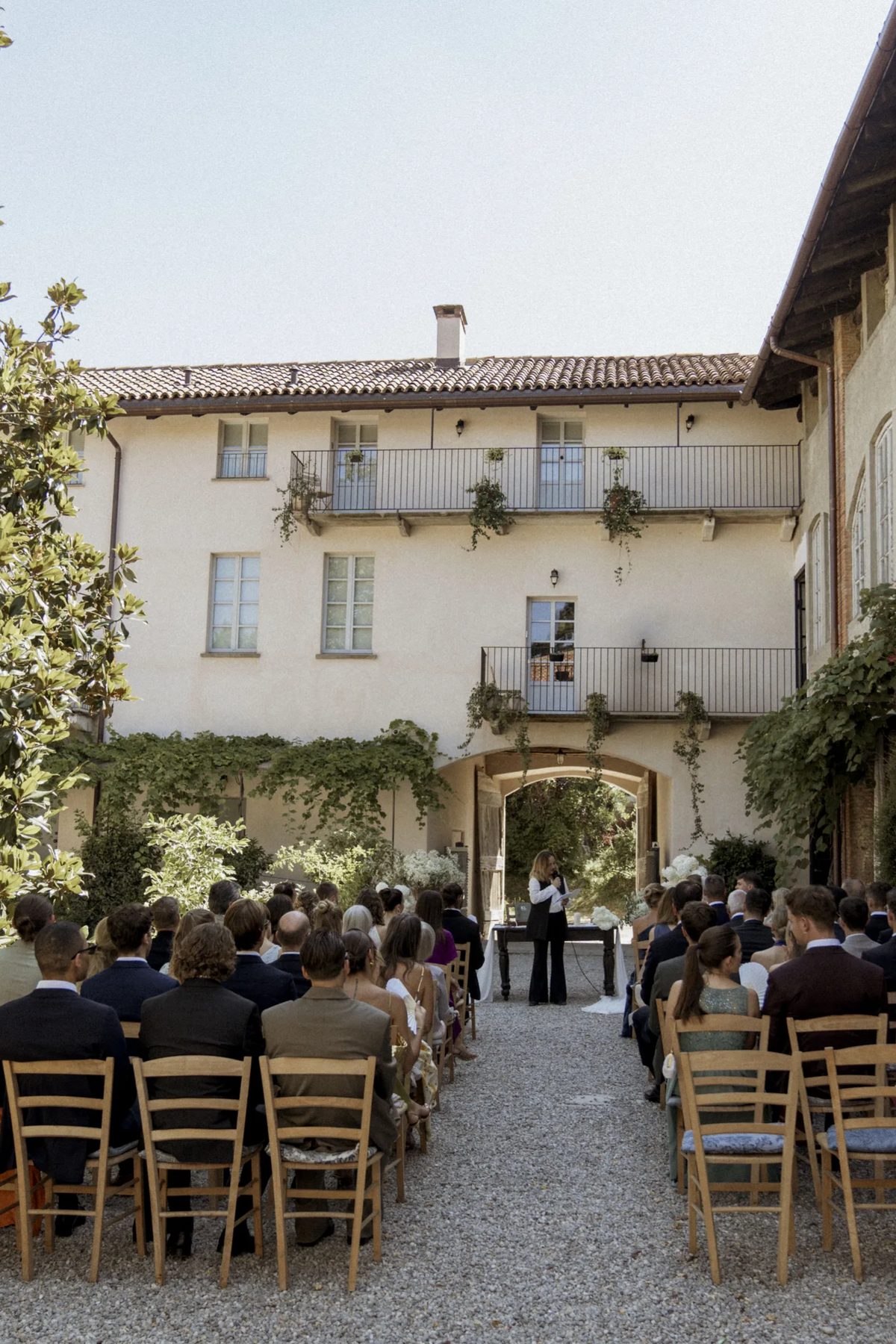 The ceremony in the gravel courtyard, seen down the aisle between guests seated on wooden chairs, with the celebrant at the far end under the old farmhouse and a magnolia in leaf